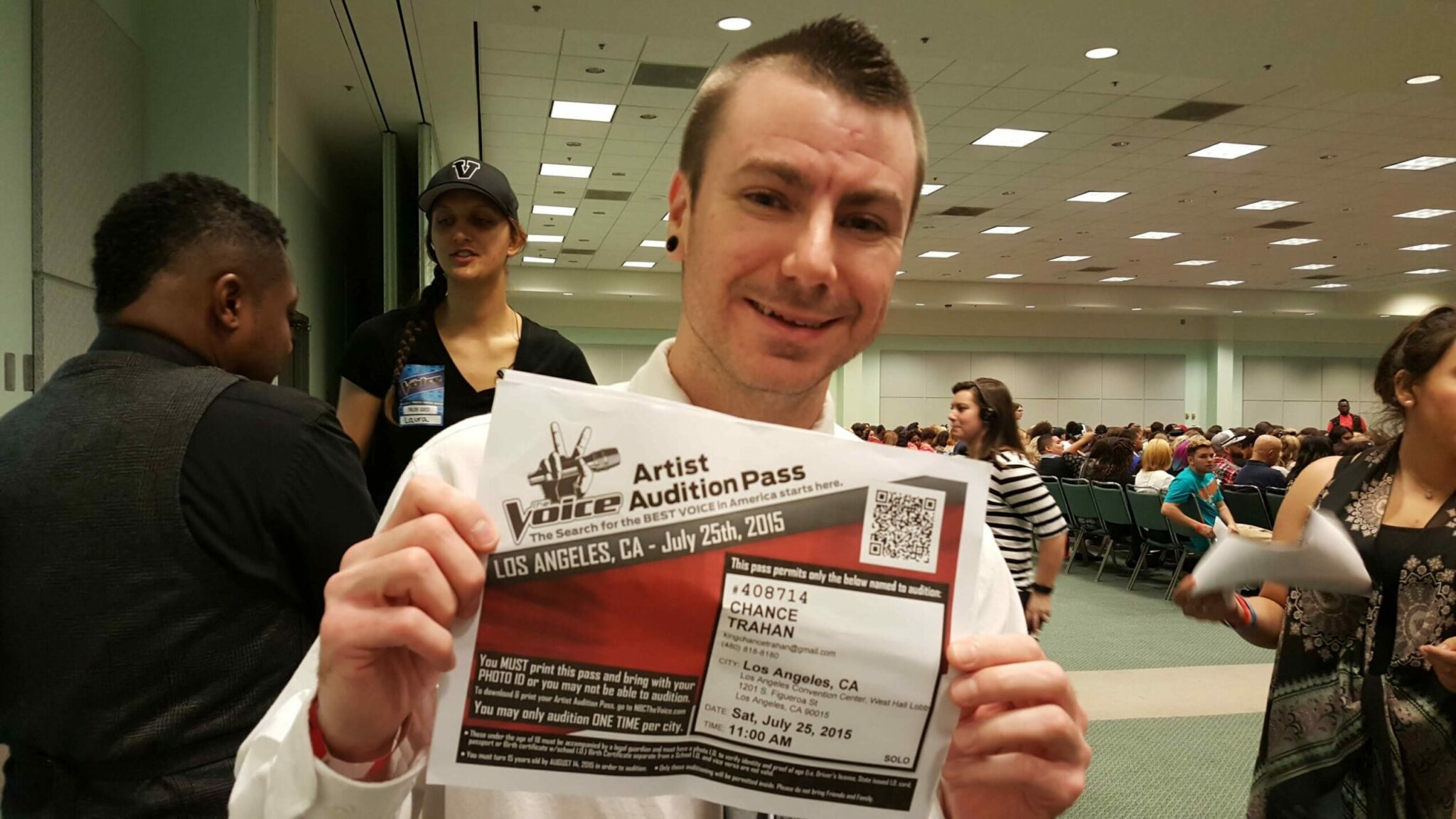 Chance Trahan holding up his audition pass for the TV show The Voice, a competing singer named Dallas Walls is in the background talking to staff